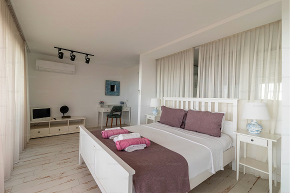 A white bedroom with two walls of curtains, one behind a white wooden bed and purple pillows, a purple blanket across the foot of the bed, and the rest in white linens. Two sets of towels are on the bed, one is pink and one is white in each set. A pair of matching bedside tables with lamps sit either side of the bed. A small white desk with a black chair is visible at the end of the room, and a white dresser with a television and fan is nearby the desk. Three black ceiling lights are angled at the desk and dresser. The flooring is light-colored hardwood.