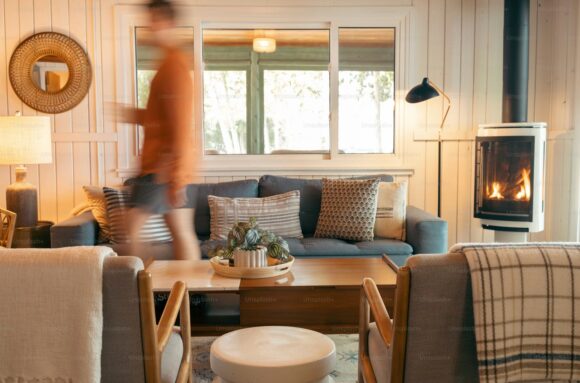 A person in motion walking through a living room with white wood panelled walls, a wood-burning fireplace in the corner, a standing lamp next to a blue two seater sofa, and two arm chairs with woven blankets on the backs. A wooden coffee table is placed between the seating, and there's a table lamp visible below a round mirror on the wall. A large window to a patio or exterior space allows bright light to stream into the room.