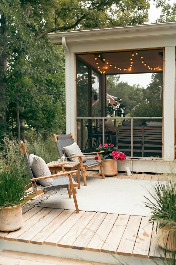 An exterior view of a tree-lined small wooden deck with 2 chairs, a side table, exterior rug, and a large potted plant with flowers. Behind is a screened porch with a dining or seating area and string lights overhead. You can see through to the green trees beyond, and a red building in the distance. Grasses surround the edges of the wooden deck, with two more large potted green plants at the front.