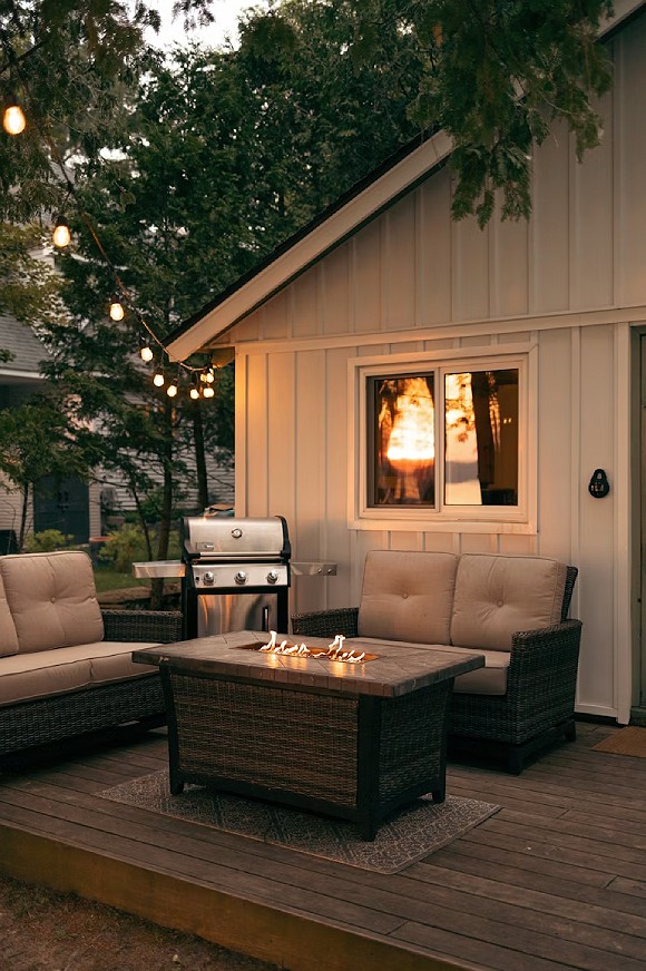 Outside a white wooden paneled building are two dark-colored outdoor loveseats with off-white cushions and a square firepit table on a small rug on the dark wood decking. In the corner sits a silver and black BBQ. String lights hang from the corner of the building into the trees, over the deck and seating.