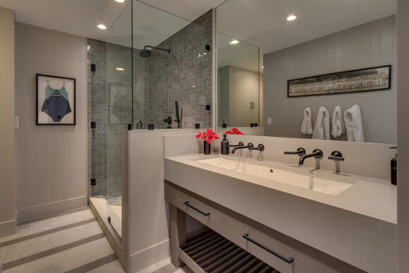 A cream and beige bathroom with grey tiling and black fixtures in the glass-walled shower, light-colored countertops that match the kitchen, and pale wood cabinetry underneath the double trough sink. Towels hang on hooks on the opposite wall, beneath a long framed piece of art, which is all visible in the large mirror. Recessed lighting shines down from the light-colored ceiling, and a framed blue and turquoise bathing suit hangs on the wall near the shower. A posey of red flowers sits in the corner of the counter by the sink.