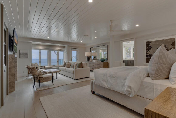 The bedroom features a white slatted wooden ceiling running towards the window at the far end with a view of the water. A seating area with a sofa and two chairs plus a side table and television has a dresser behind the sofa, all of which is near the view. Towards the front right, the bed is dressed in white linens with an oversize tan pillow and a medium-brown wood bedside table. Beyond the bed, a map appears on the wall next to an open doorway.