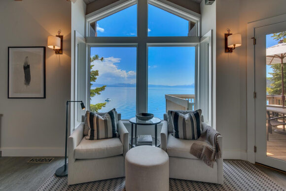 Two armchairs with brown and black striped pillows against the backs and a blanket over the arm of one sit with a small glass table between them, and a round footstool in front. Behind the chairs is a floor-to-ceiling arched window to the bright blue lake and sky outside. Two wall sconces either side of the window provide additional light. A tan and black checkered rug grounds the seating area. A framed piece of art hangs on the wall to the left, while to the right is a door to an exterior deck with an umbrella, table and seating.