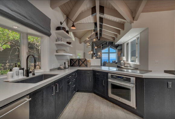 A kitchen with no visible upper cabinets, lower cabinets in dark grey, appliances in stainless steel, and light grey countertops contrasts with the light tan exposed beam vaulted ceiling. A window to the left shows greenery with a grey roman blind pulled up to expose the view. Open shelves are on one wall with a wall sconce above that extends out and is adjustable. A large window at the back shows the blue sky and water.