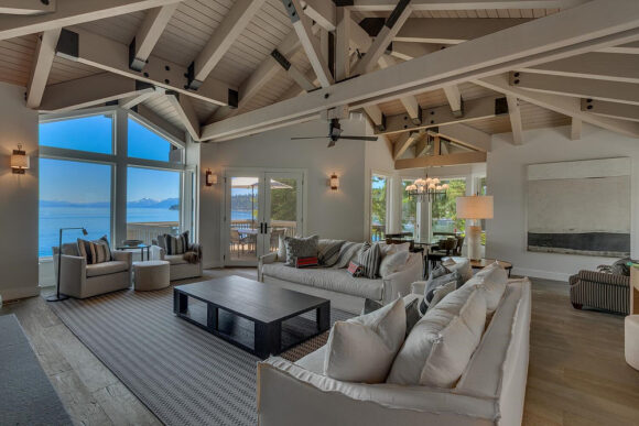 A neutral light-colored beige, tan, brown, and grey living room with beige exposed-beam vaulted ceilings, two cream sofas at a 90-degree angle, a dark wooden coffee table in front of them. There's a smaller seating area in front of the window with a view to the lake, and behind the sofas there's another smaller dark striped sofa and seating area near some art. The hardwood flooring is medium brown, and a grey striped rug anchors the bigger sofas and chairs. Lighting is cast by wall sconces and table lamps scattered through the space, as well as the large window with a view to the lake and blue sky.