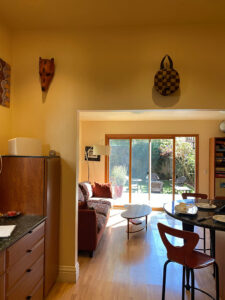 BEFORE showing the kitchen wall and passageway to the dining room, painted a bright yellow. A large medium-colored wood cabinet is next to the wall, and a dark marble counter top is visible above drawers. On the right, the kitchen counter is rounded, with a view through the living room (sofa on the left) to the backyard patio, and green space with seating and plants. Collectible masks hang on the wall above the opening.