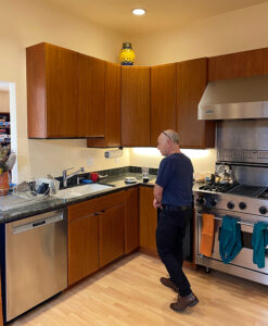 BEFORE in the kitchen, looking towards the corner with the sink and dishwasher, and cherry wood upper and lower cabinets. The upper cabs don't extend to the ceiling, gathering dust and oils from cooking. The flooring is a light oak finish hardwood. A large oven and cooktop in stainless steel is visible with a kettle on the surface.