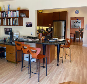 Kitchen before the changes was mismatched, dark marble pill-shaped counter end with black legs, a fridge behind, flooring running front to back, and some counter-height chairs with orangey wood backs and seats on black legs. Lots of paperwork and books strewn around, and an open shelf on the wall to the left full of books.