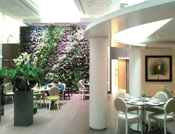 A restaurant with multiple seating areas. White chairs with beige circle patterned fabric seats and backs surround round and square glass tables. A white column and white partial soffit are visible overhead of one table. To the left there is a large planter on a square cement stand, and beyond that is another dining area, with a wall of multi-colored plants at the very back. Bright sunlight streams in from a skylight above. A woman sits at one of the tables, reading.