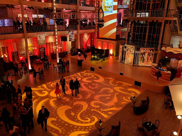 The San Francisco Design Center central event space, lit up in orange, red, and yellow lights, including paisley patterns created on the floor with yellow and orange lights. People are walking across the floor as well as gathering around the edges of the room at tables and displays.