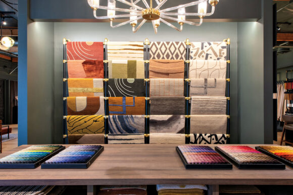 A fabric showroom featuring multiple patterns and colors of fabric on stretchers, with additional colors and fabrics in trays on a wooden table in front of the display wall. A chandelier overhead provides bright light.