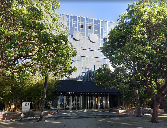 The exterior of the San Francisco Design Center Galleria building, surrounded by trees, with a walkway and railing leading to the multiple glass door entrance. Above the door is a black awning and above that are more levels of glass windows with two circles at the top containing some of the letters that spell out SFDC.