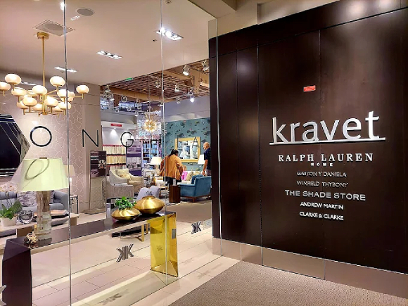 The entrance to Dongia and Kravet showrooms, with two people in the background looking at furniture. Towards the front are lamps, chandeliers, side tables, sofas, and a gold and black console table. The Kravet logo wall is black with white writing, and the Dongia logo is black on clear glass.